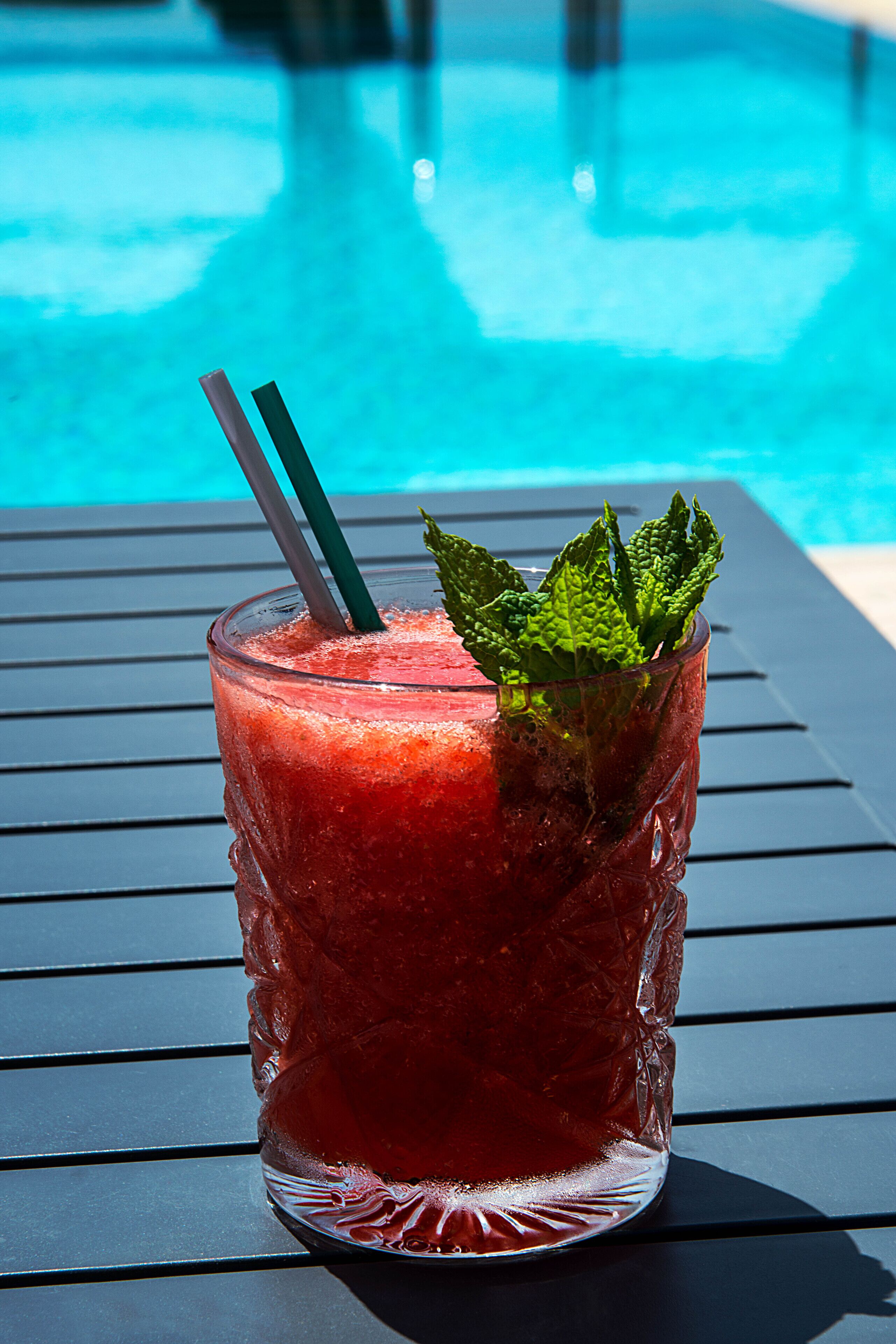 Un vaso de bebida roja con una ramita de menta y dos pajitas sobre una mesa de metal junto a una piscina.