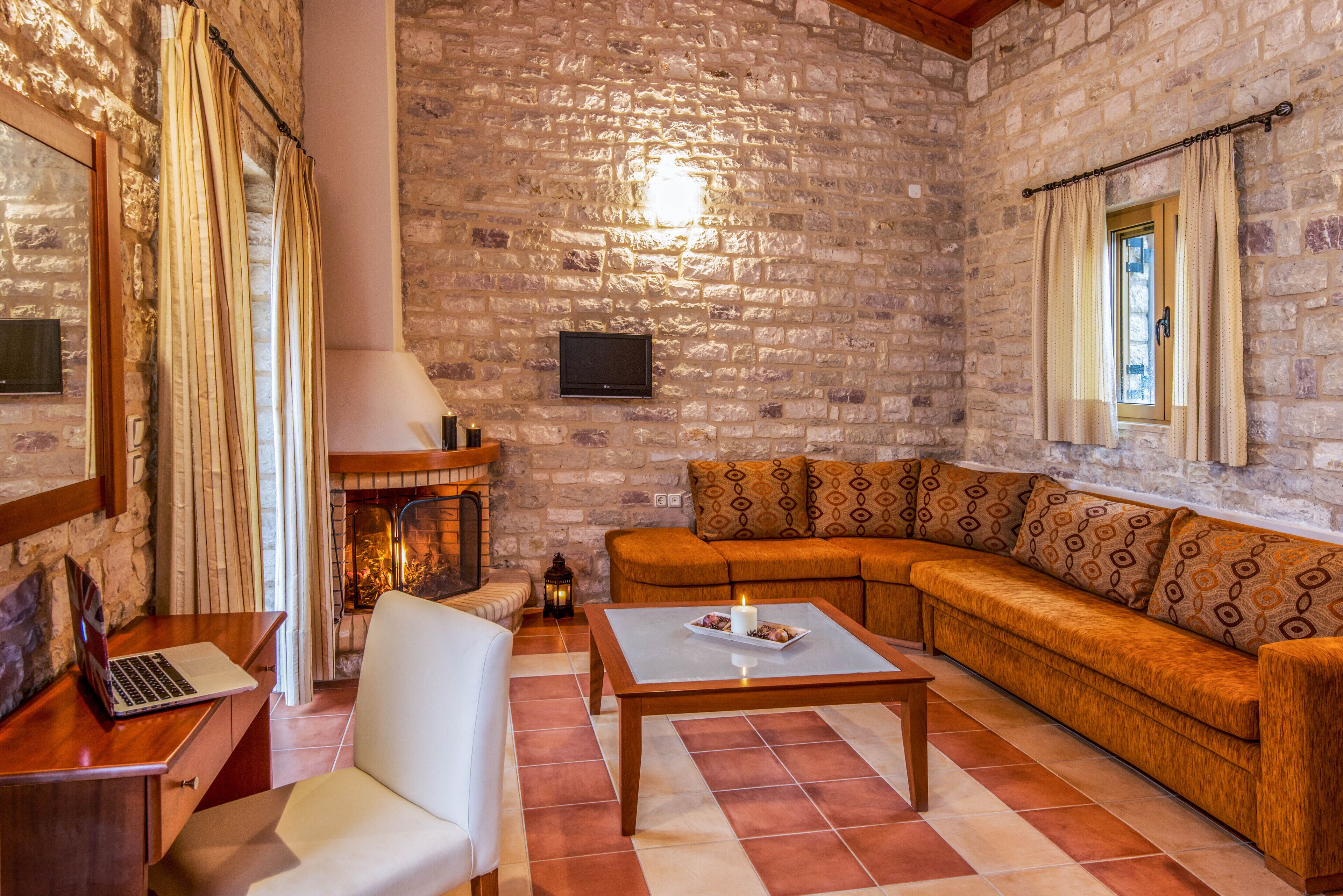 A cozy living room with a stone wall, a fireplace, a sofa, a coffee table, and a laptop on a wooden table.