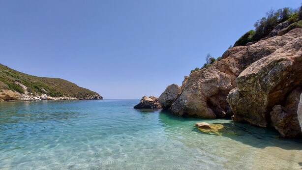 A coastal landscape with clear turquoise water, rocky formations, and a hilly backdrop. - Seagulls - Within walking distance from sea! (Agioi Apostoloi)