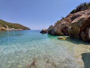 A coastal landscape with clear turquoise water, rocky formations, and a hilly backdrop. - Seagulls - Within walking distance from sea! (Agioi Apostoloi)