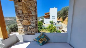 A balcony with a white cushioned bench, a stone pillar, and a view of a white building and greenery in the background. - Seagulls - Within walking distance from sea! (Agioi Apostoloi)