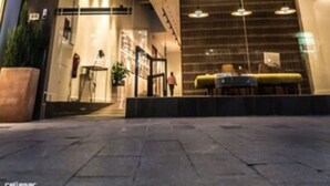 Entrada de un hotel con diseño moderno, destacando un frente de vidrio, una recepción y una escalera que lleva a los pisos superiores.