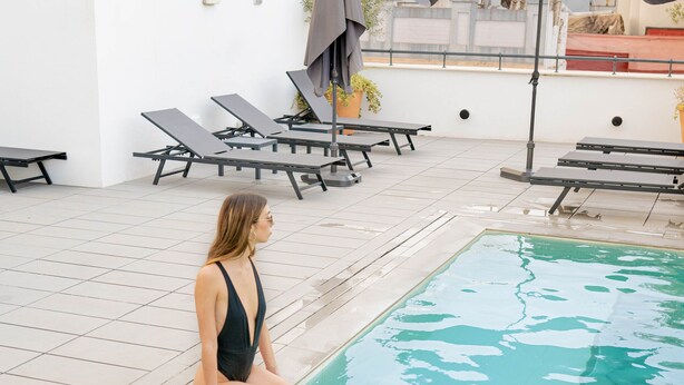 Una mujer sentada junto a una piscina, con sillones de descanso y sombrillas al fondo.