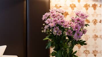 Un lit avec un oreiller blanc, une petite table ronde ornée d’un vase de fleurs violettes et un mur décoré d’un motif floral.