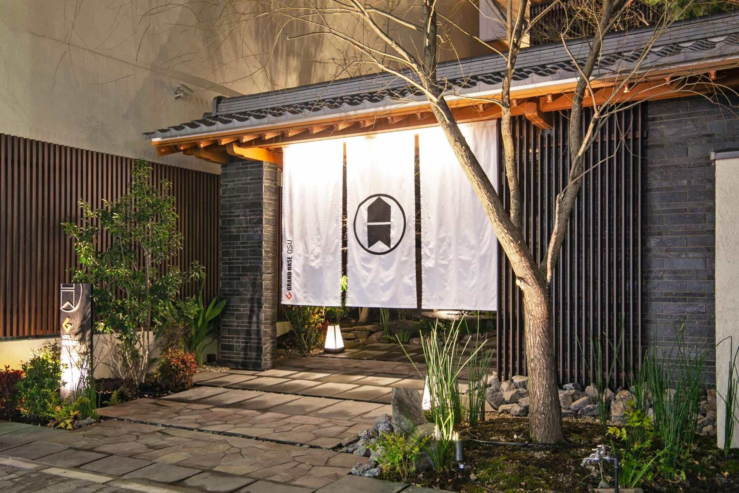 Un edificio de estilo japonés tradicional con entrada de madera, un camino de piedra y un jardín bien cuidado con plantas y un árbol.