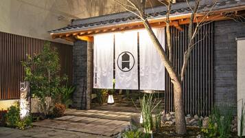 A traditional Japanese-style building with a wooden entrance, a stone pathway, and a well-maintained garden with plants and a tree.