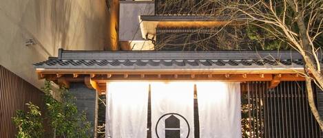 A traditional Japanese-style entrance with a tiled roof, a white banner with a black emblem, and a stone pathway leading to the entrance.