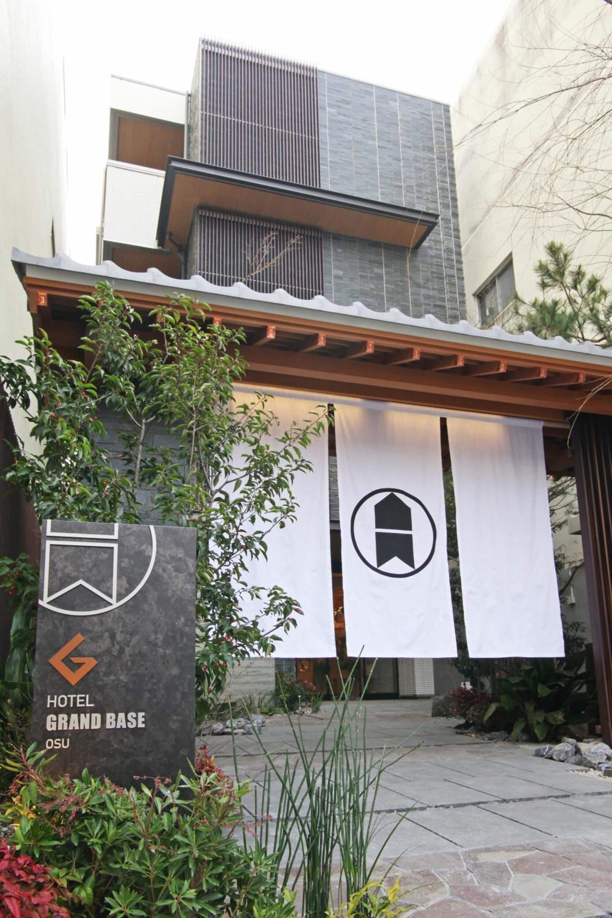 A hotel entrance with a modern facade, a sign indicating 'Hotel Grand Base OSU', and a covered walkway with a pergola.