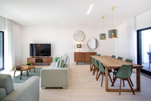 A modern living room with a flat-screen TV, a sofa, a dining table with chairs, and a wooden cabinet.