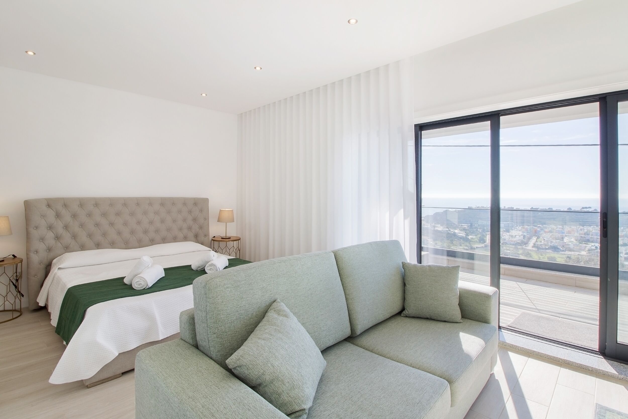 Et moderne hotelværelse med en stor seng, en sofa og udsigt over byen gennem gulv til loft-vinduer.