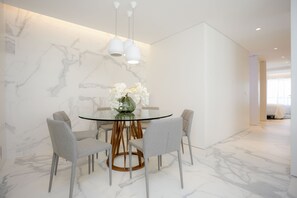 A modern dining area with a glass table, grey chairs, a vase with white flowers, and a marble wall.