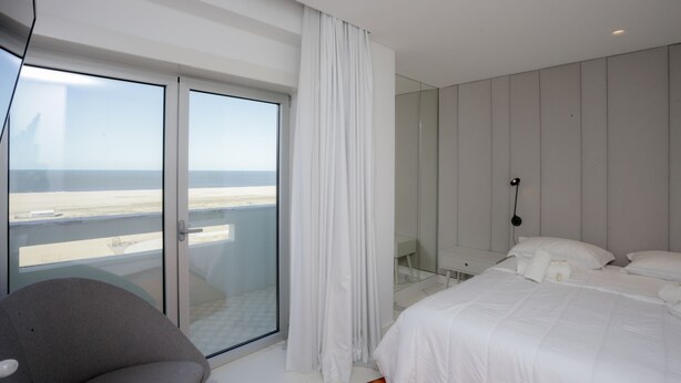 Ein modernes Hotelzimmer mit einer großen Glasschiebetür zum Strand, einem Bett mit weißen Leinentüchern, einem grauen Sessel und einem Nachttisch mit einer Lampe.