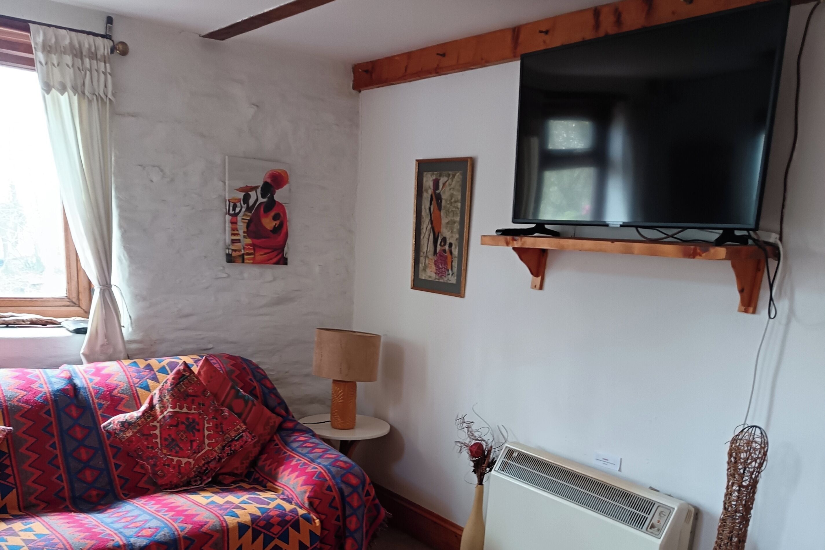 A living room with a sofa, a television mounted on a wooden beam, a wall-mounted air conditioner, and a window with curtains.