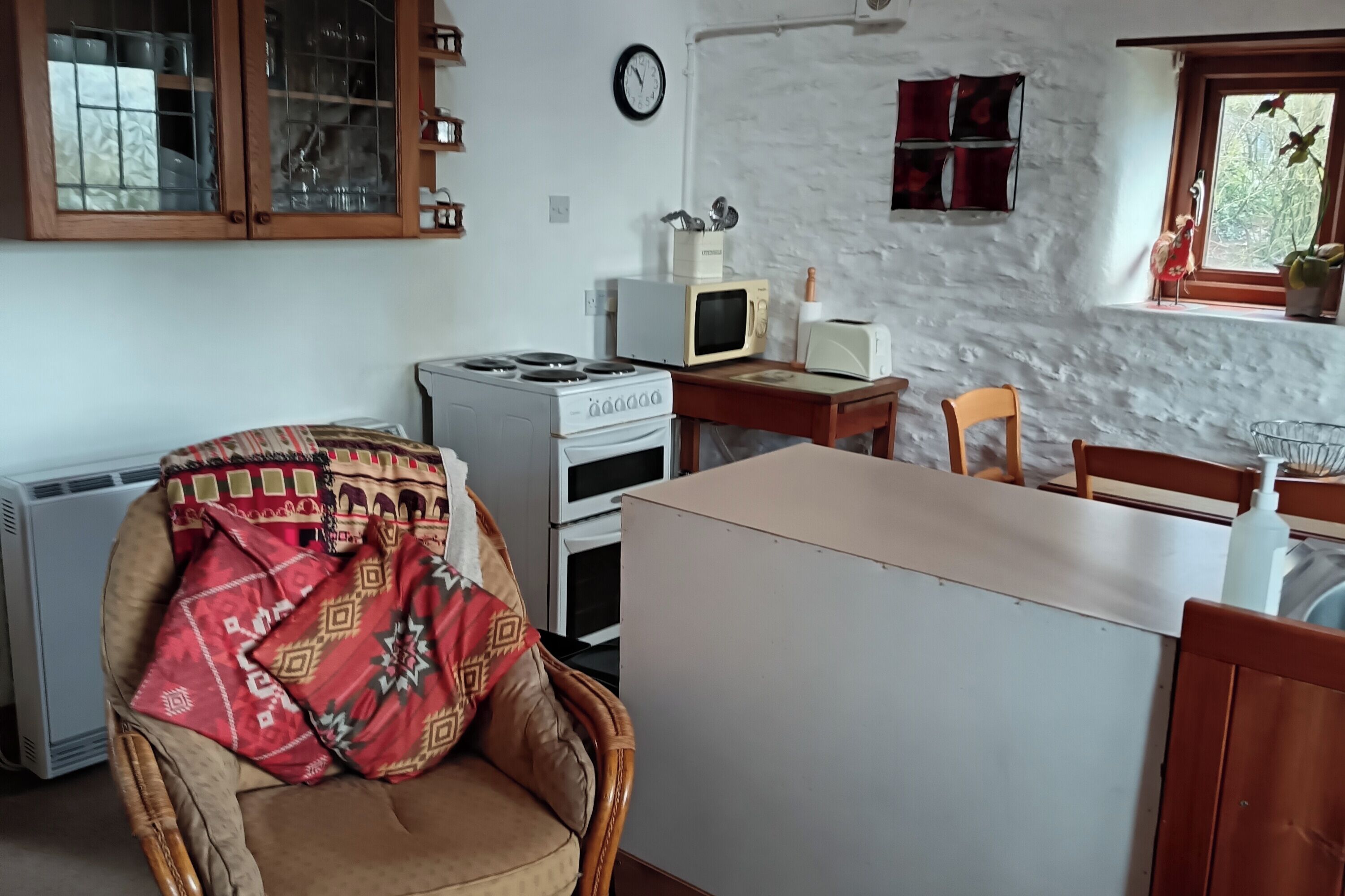 A compact kitchen with a stove, microwave, and toaster. There is a chair with a patterned cushion, a wooden table, and a window with a view of greenery.