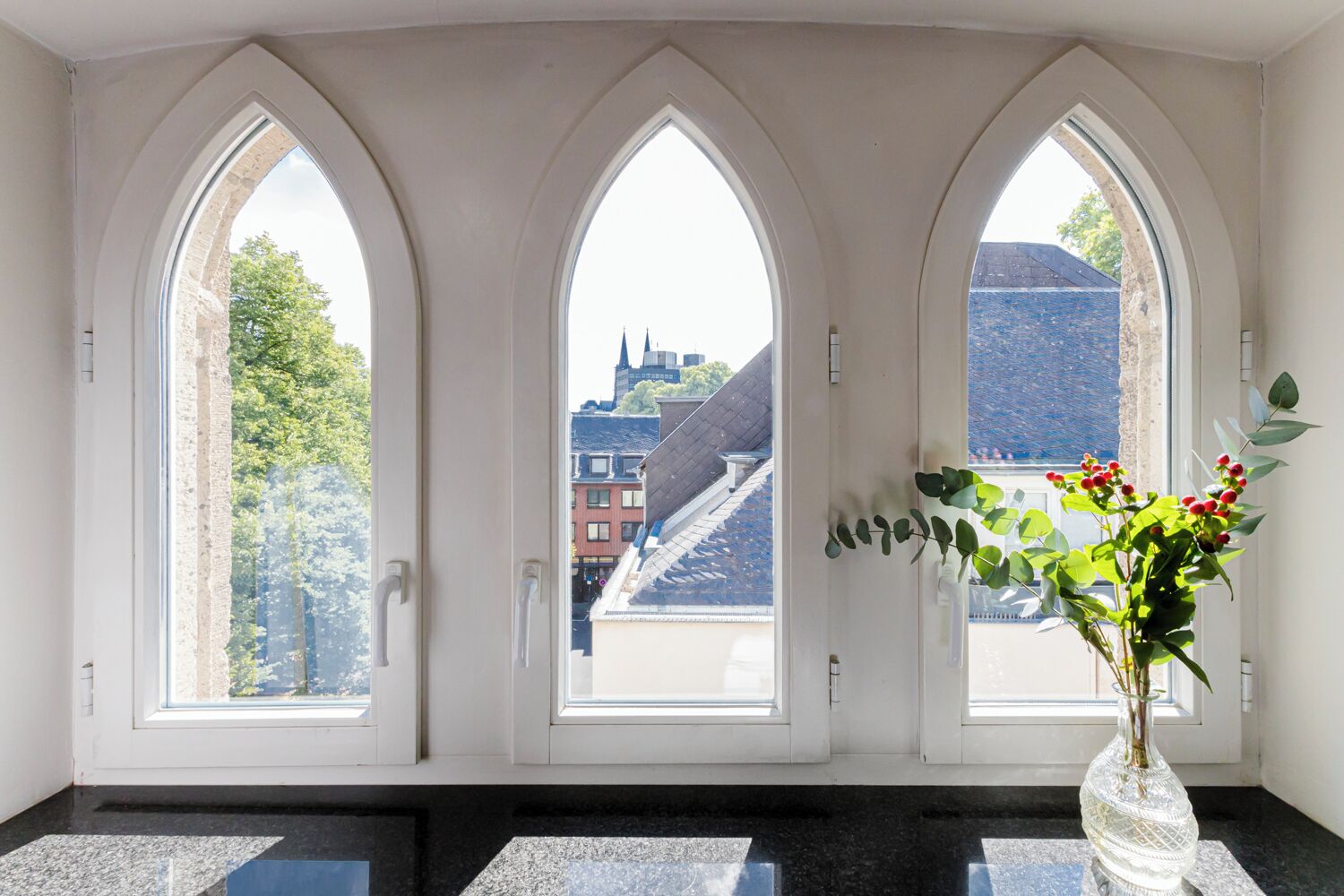 A room with three arched windows, a vase with flowers, and a view of buildings and trees.