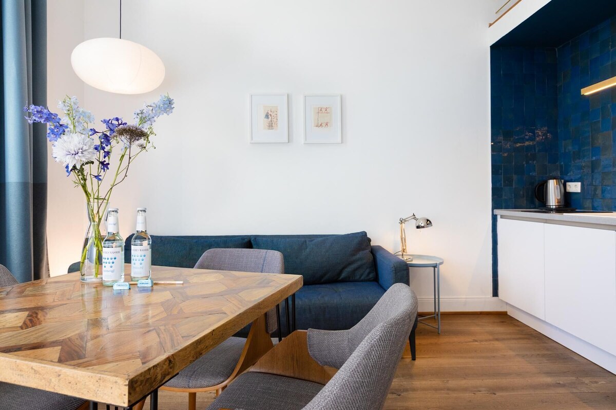 A modern living room with a wooden dining table, grey sofas, a blue accent wall, and a kitchen area with a blue backsplash.