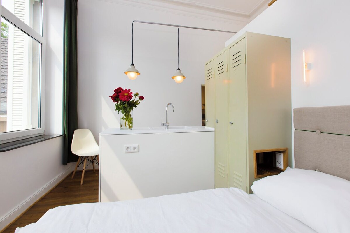 A hotel room with a bed, a small desk with a sink, a chair, and a wardrobe.