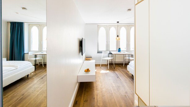 Ein modernes Hotelzimmer mit Holzboden, einem weißen Bett, einem kleinen weißen Tisch mit Obstschale, einem an der Wand montierten Fernseher und einem Essbereich mit Tisch und Stühlen.