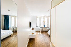 Una habitación de hotel moderna con suelo de madera, una cama blanca, una mesita blanca con un frutero, un televisor montado en la pared y una zona de comedor con mesa y sillas.
