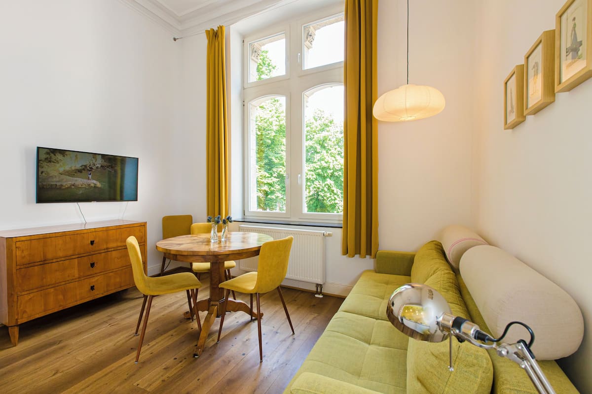 Une pièce comprenant une table à manger, des chaises, un canapé, un radiateur, une fenêtre avec des rideaux et un tableau accroché au mur.