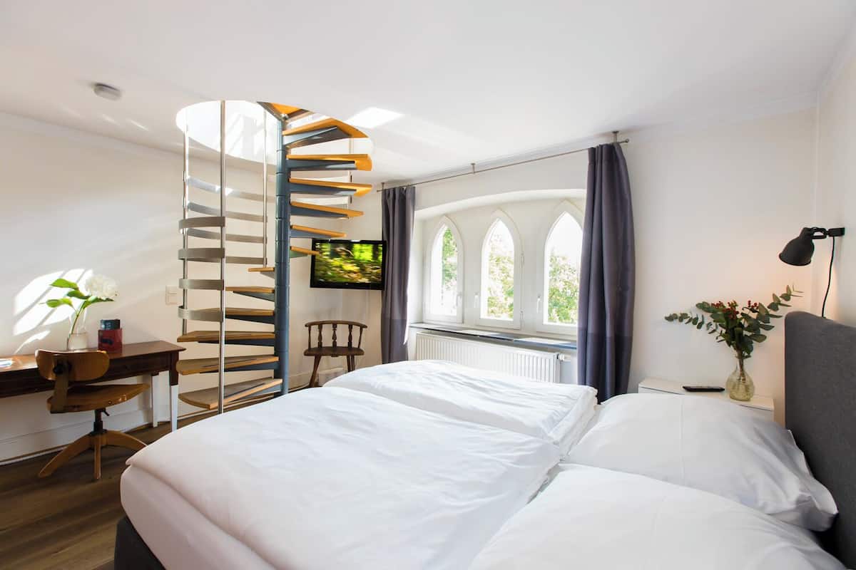 Une chambre d’hôtel moderne avec un escalier en colimaçon, un lit avec du linge de maison blanc, un bureau avec une chaise, une télévision et une fenêtre donnant sur un paysage verdoyant.