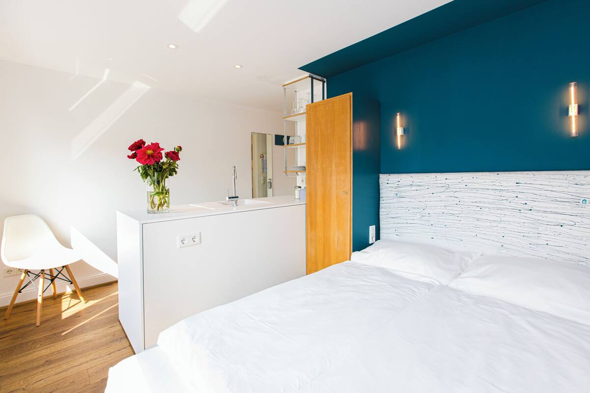 Une chambre d’hôtel moderne avec un lit, une chaise blanche, un meuble en bois et un vase rempli de fleurs rouges.