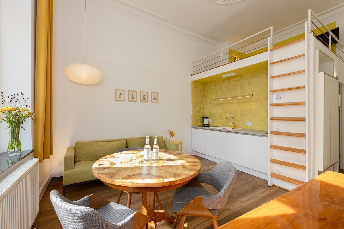 A modern living space with a round wooden dining table, a sofa, and a kitchen area with a yellow backsplash.