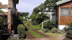 A narrow, paved pathway leading to a residential area with brick houses, lush greenery, and a wooden structure with a metal gate. - Apartment for 2 people  (La riviere saint louis)
