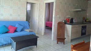 A living room with a blue sofa, a small black table, and a wooden chair. A kitchen area is visible with a countertop, a coffee maker, and cabinets. - Apartment for 2 people  (La riviere saint louis)