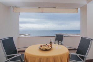 Une table ronde recouverte d’une nappe jaune, deux chaises, un panier de fruits, deux verres à vin et une bouteille, le tout sur un balcon donnant sur une plage.