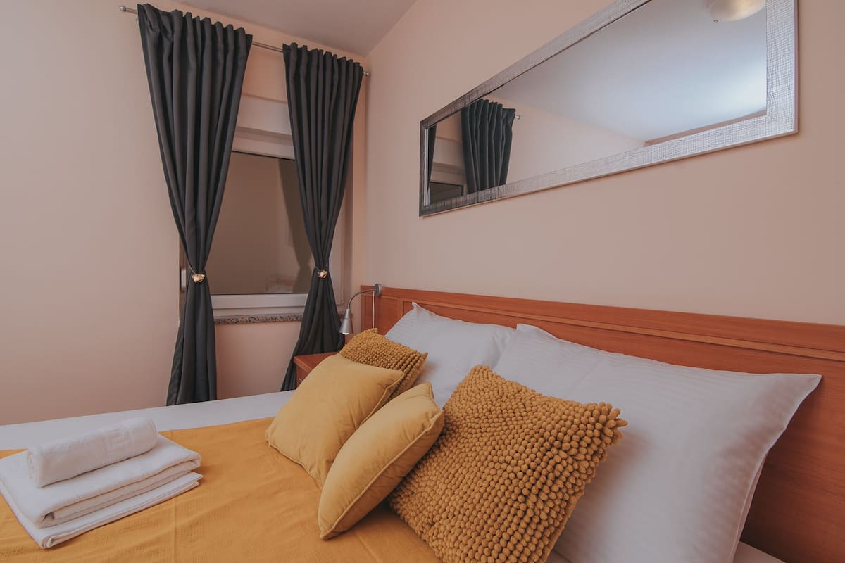 Una cama bien hecha con cubrecama blanco y color mostaza, dos almohadas color mostaza y una toalla blanca doblada. Un espejo grande cuelga sobre la cama, reflejando las cortinas y parte de la habitación.