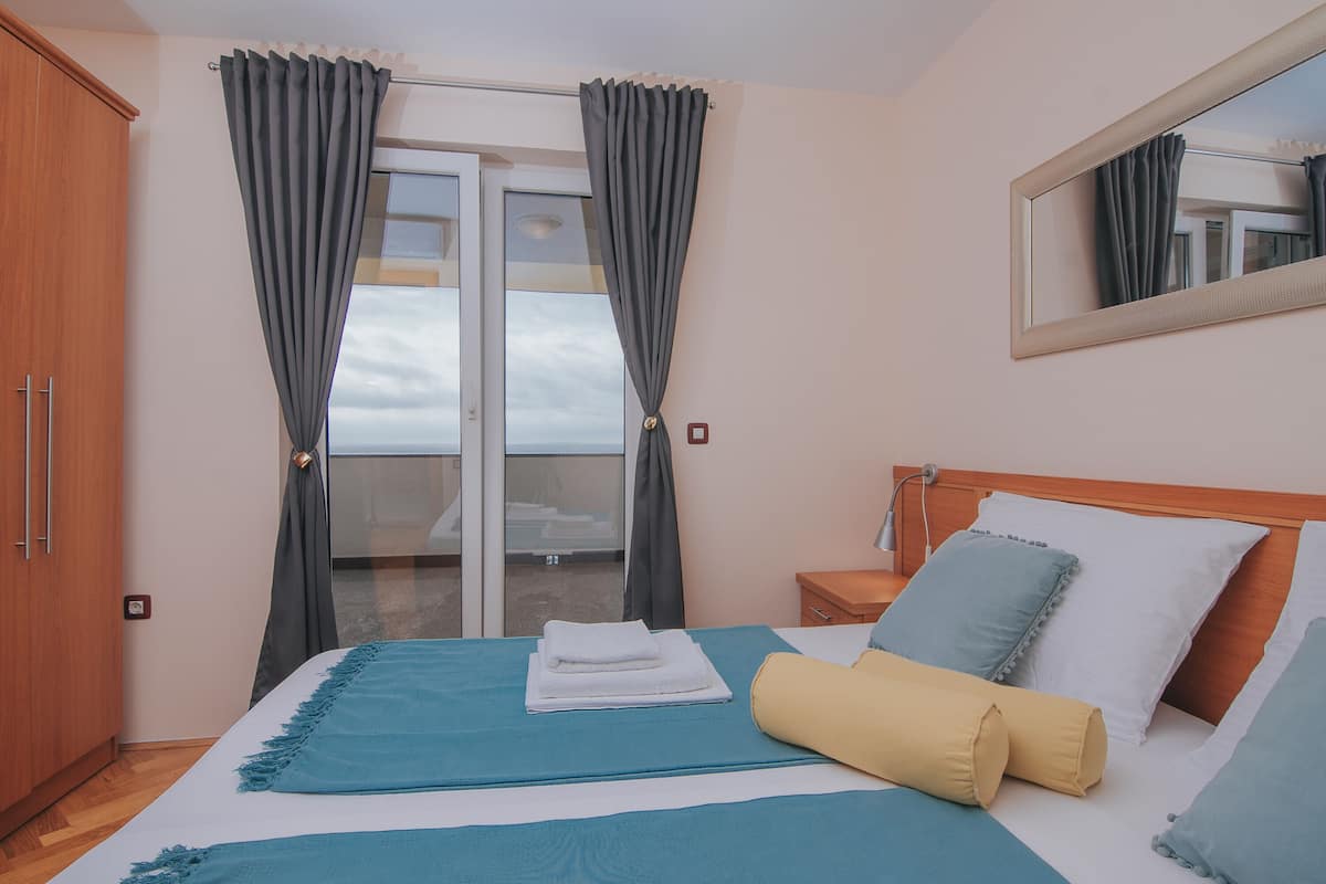 Una habitación de hotel con cama, armario de madera, mesita de noche, espejo y balcón con cortinas.