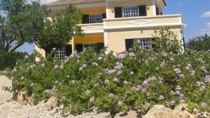 A two-story house with a tiled roof, a balcony, and a garden with purple flowers. - Casa dos Ventos (Olhao)