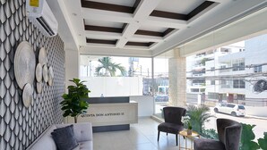 Un espace d'accueil moderne avec un comptoir de réception blanc, un canapé gris et deux fauteuils. Le comptoir de réception porte une enseigne indiquant « Quinta Don Antonio II ». De grandes fenêtres offrent une vue sur la rue et les bâtiments environnants.