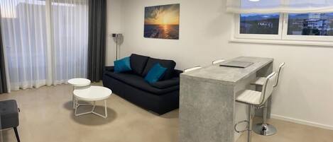 A modern living room with a sofa, two round tables, a bar area with stools, a large window with a city view, and a painting of a sunset.