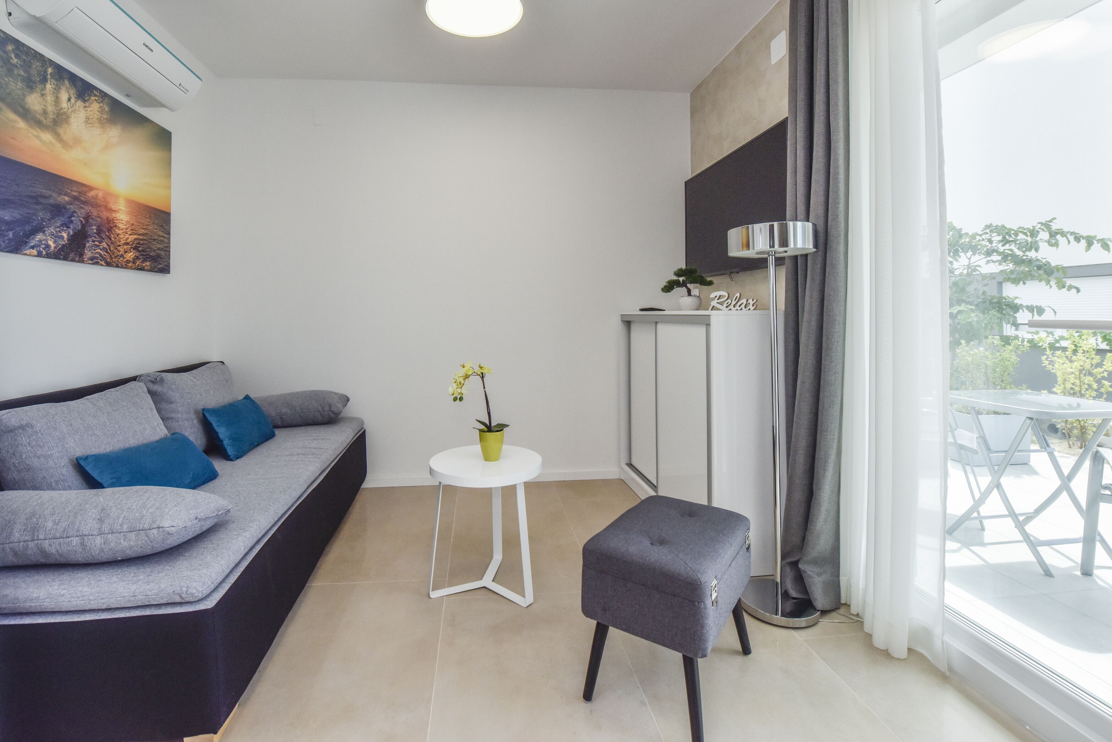 A modern living room with a grey sofa, a small white table with a potted plant, and a grey ottoman.