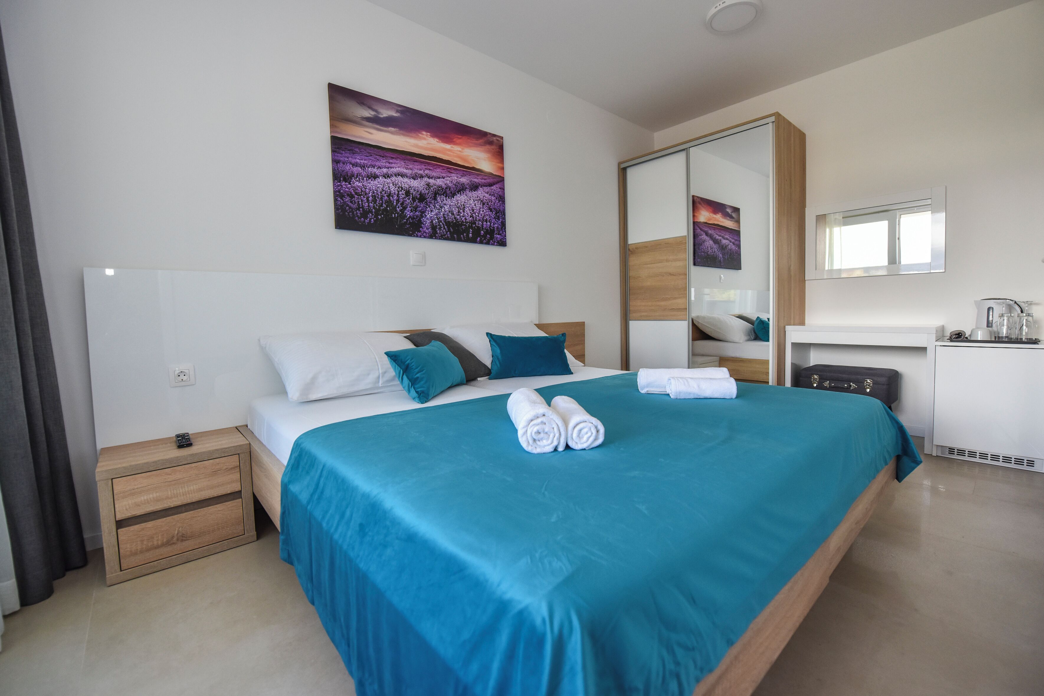 A neatly made bed with a blue bedspread, two rolled towels, and a nightstand with a remote control.