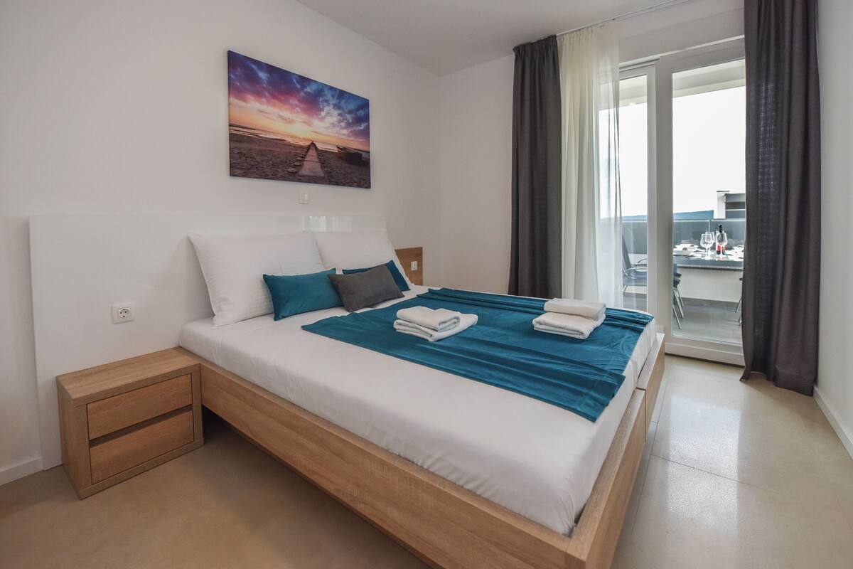 Een moderne hotelkamer met een groot bed, een nachtkastje, een schilderij van een zonsondergang en uitzicht op het balkon.