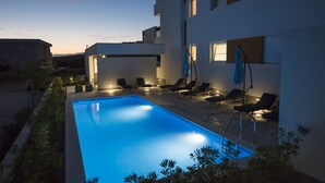 A modern outdoor pool area with lounge chairs and a well-lit pool at dusk. - Pool Apartments & Rooms Mare (Novalja)