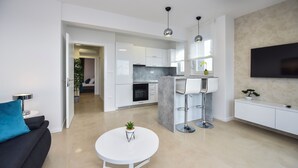 A modern living room with a white sofa, a round coffee table with a plant, and a kitchen area with white cabinets and a TV mounted on the wall. - Pool Apartments & Rooms Mare (Novalja)