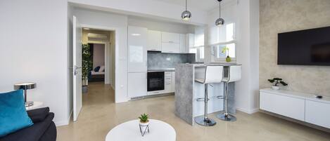 A modern living room with a white sofa, a round coffee table with a plant, and a kitchen area with white cabinets and a TV mounted on the wall.