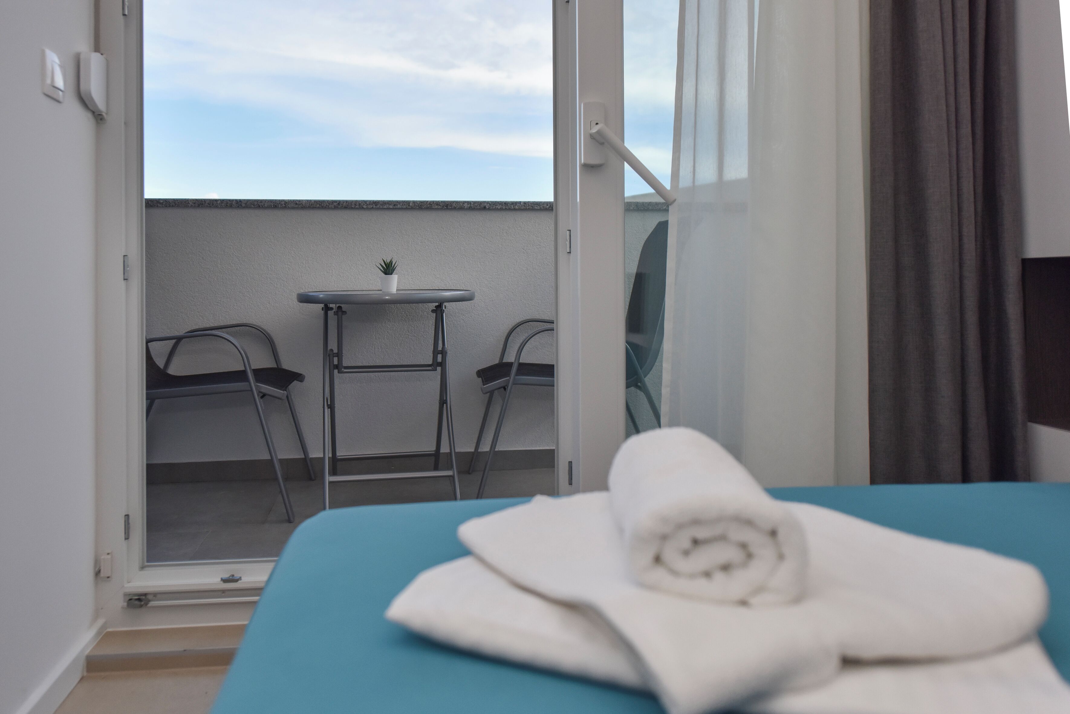 A hotel room with a bed, a small table, two chairs, and a view of the sky through an open door.