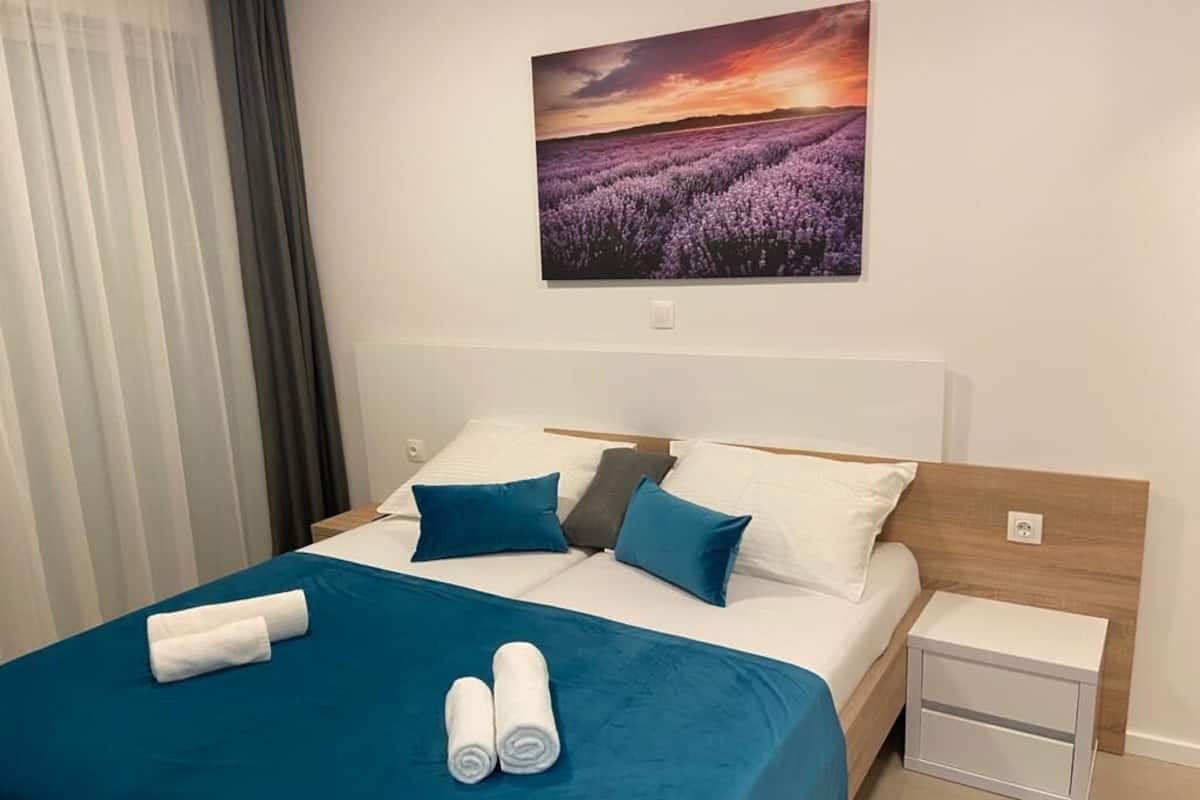 Een net opgemaakt bed met blauw-witte beddengoed, een klein nachtkastje en een schilderij van lavendelvelden aan de muur.