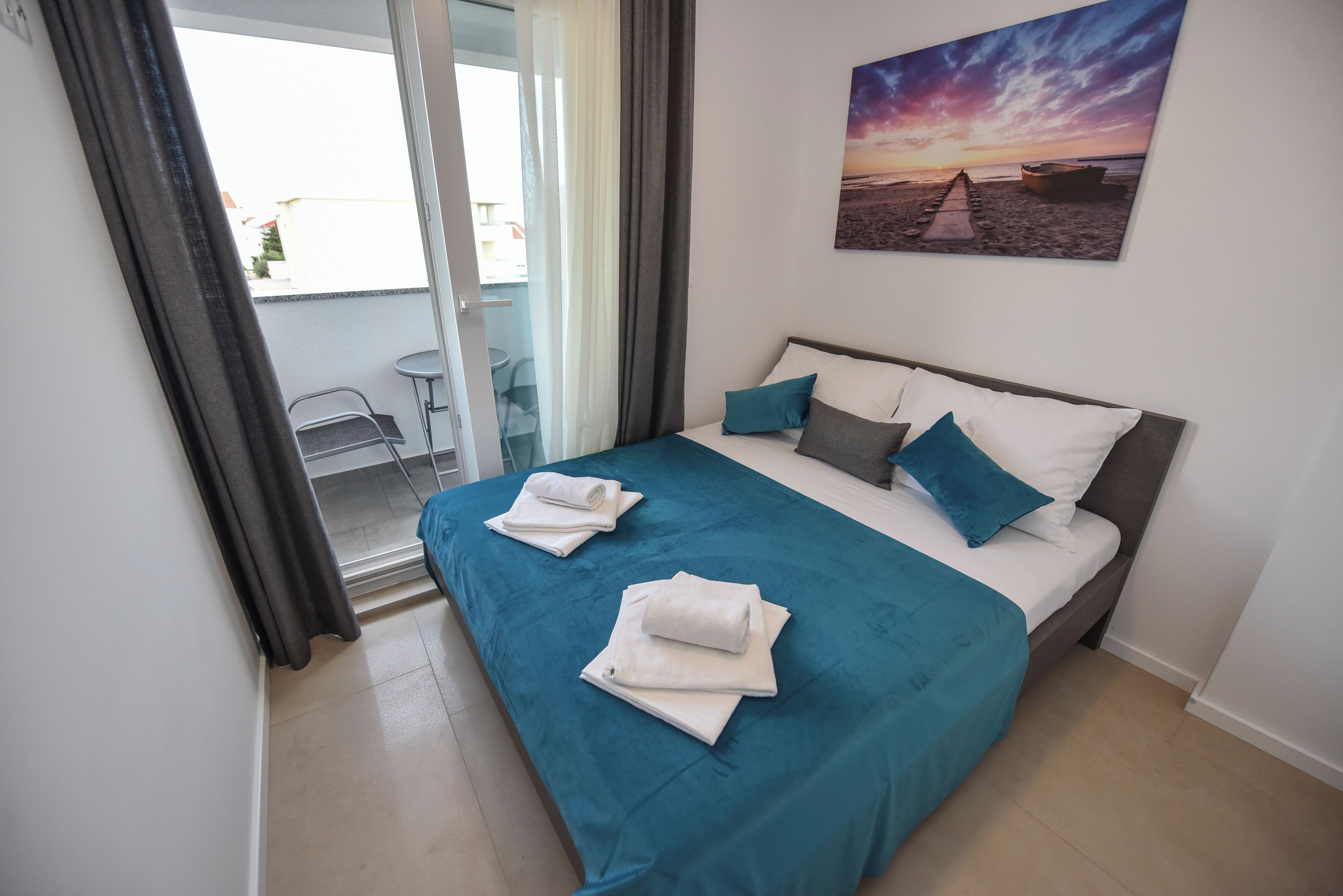 Una cama bien hecha con una colcha verde aguamarina y almohadas blancas, un cuadro de una puesta de sol en la pared, y un balcón con una silla y una mesa.