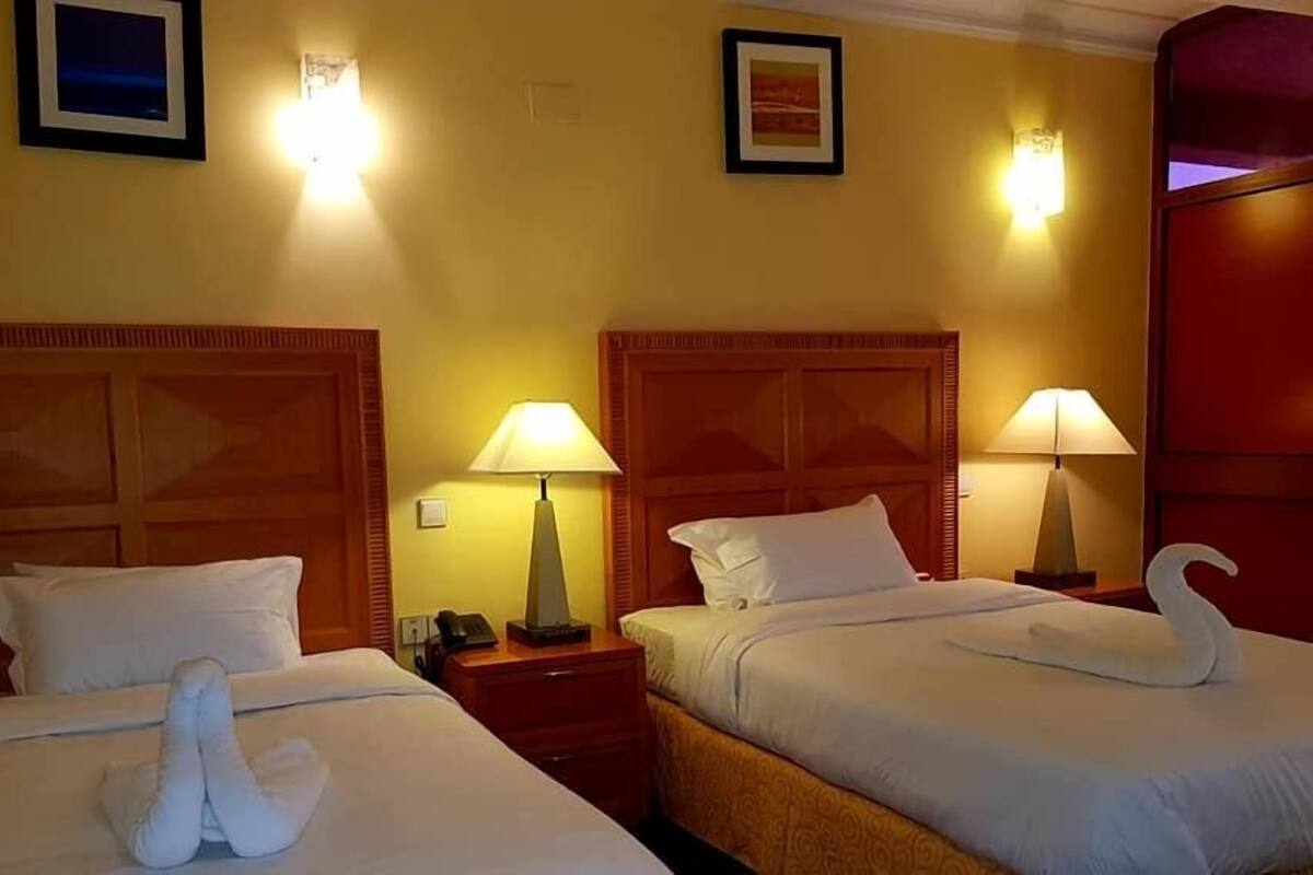 Ein Hotelzimmer mit zwei Betten, jeweils mit einem hölzernen Kopfstück, einer Nachttischlampe und einem gerahmten Bild an der Wand.