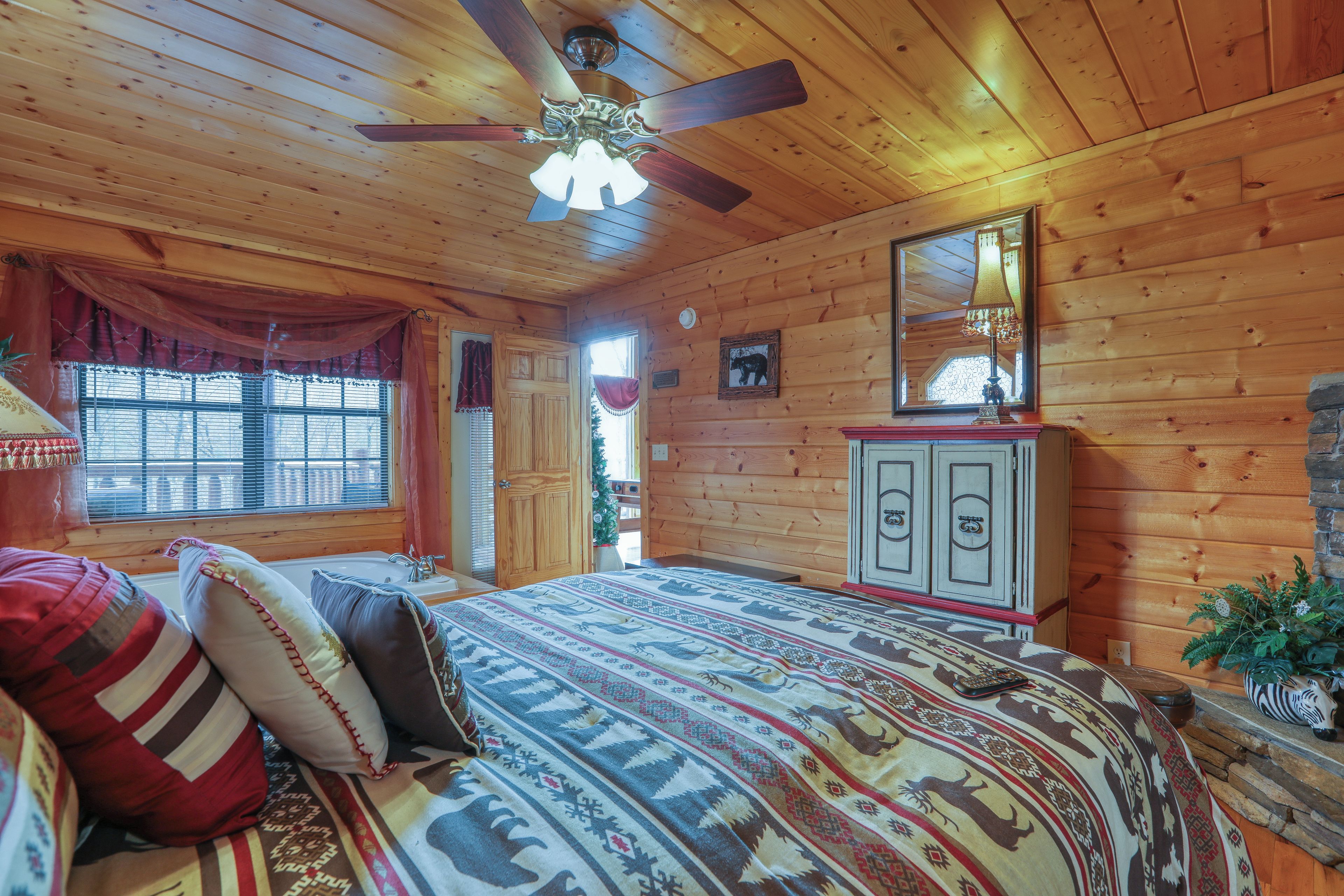 A wooden cabin bedroom with a bed, a ceiling fan, a dresser, and a window with curtains.