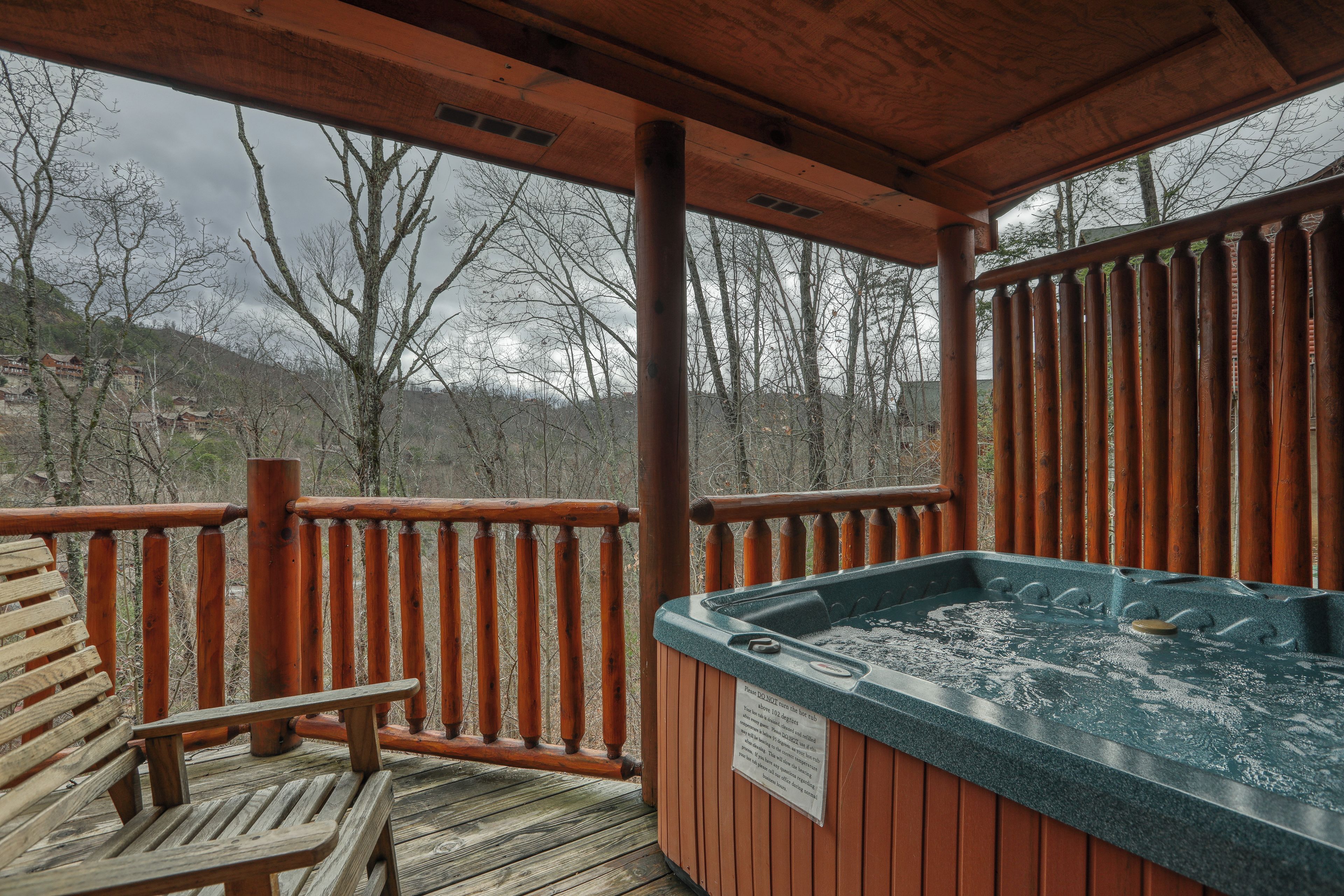 A wooden deck with a hot tub, a wooden bench, and a view of trees and hills.