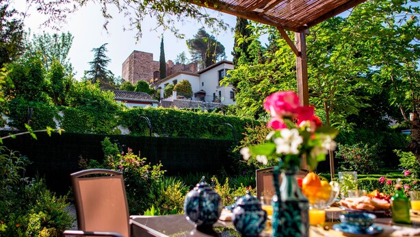 A garden patio with a table set for breakfast, surrounded by lush greenery and a view of a historic building.