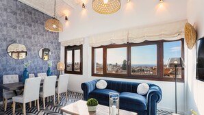 A modern living room with a blue sofa, dining table, and chairs, large windows with a city view, and decorative lighting.
