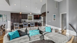 A modern living room with a grey sectional sofa, wooden dining table, and a kitchen in the background.